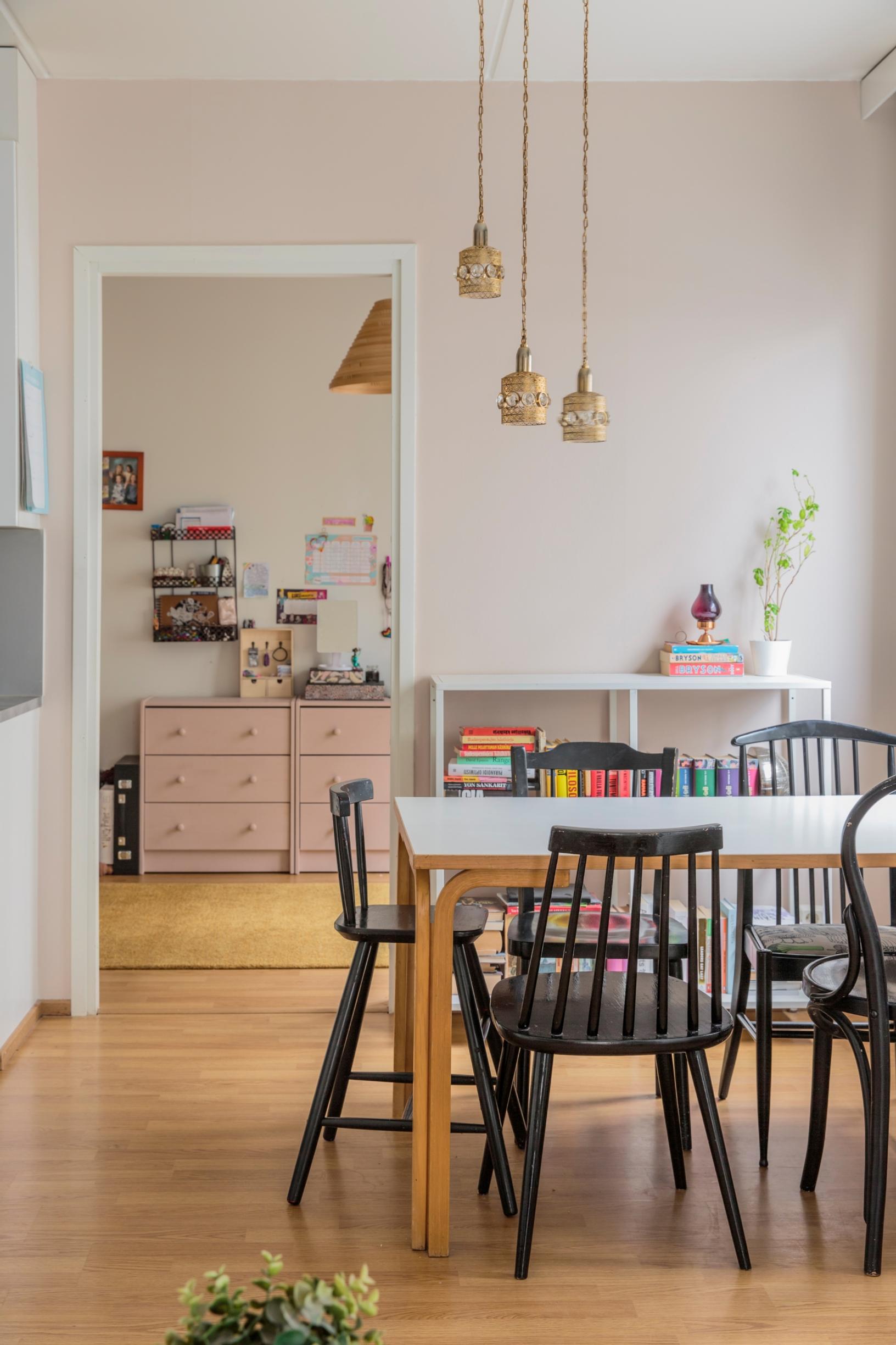 Keittiössä on ruokapöytä, jonka ympärillä on mustia ruokailutuoleja. Taustalla oviaukosta näkyy vaaleanpunainen lipasto.