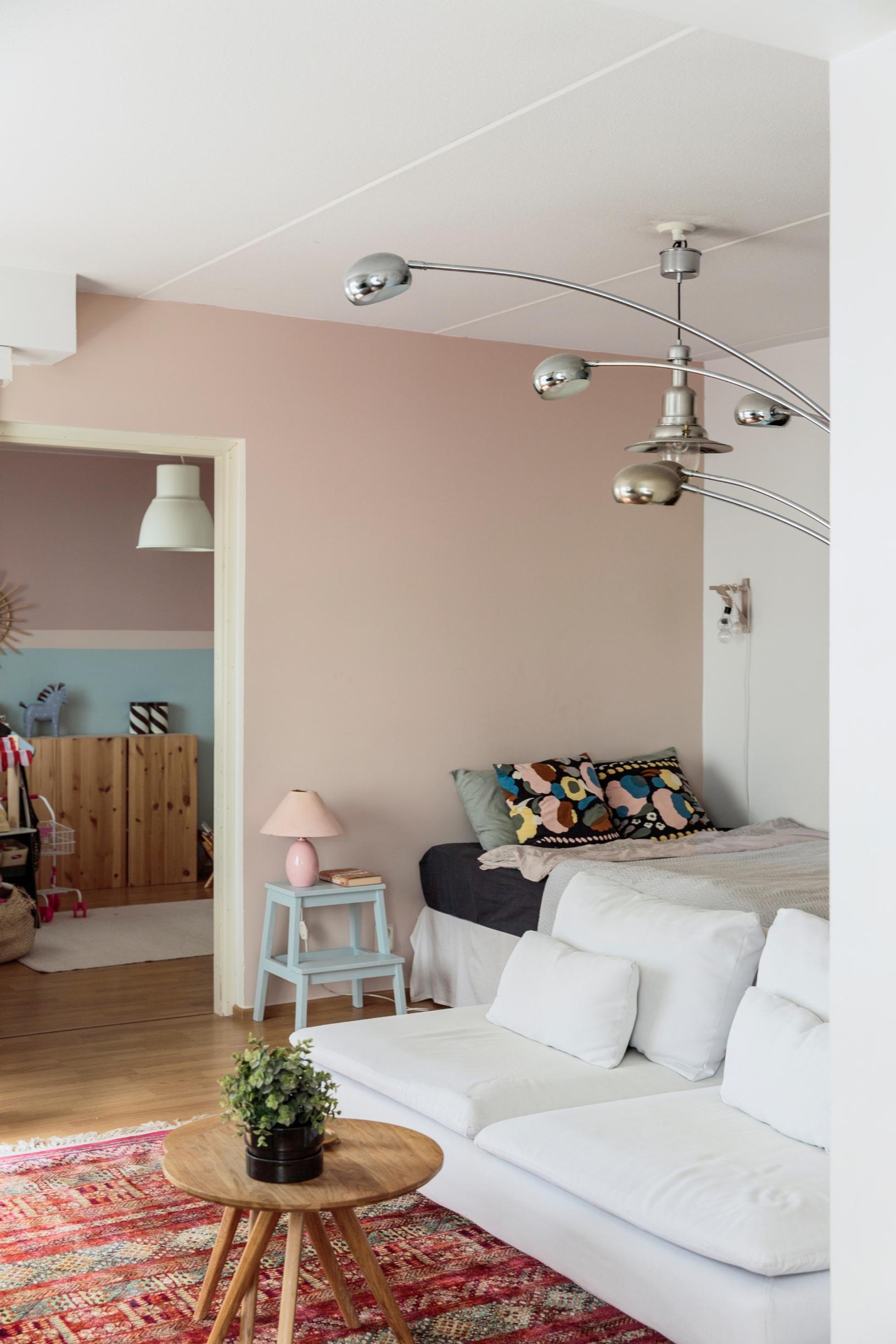 Olohuoneessa on valkoinen sohva ja värikäs matto. Taustalla näkyy sänky. Vaaleanpunainen lamppu on jakkaralla, joka toimii yöpöytänä.