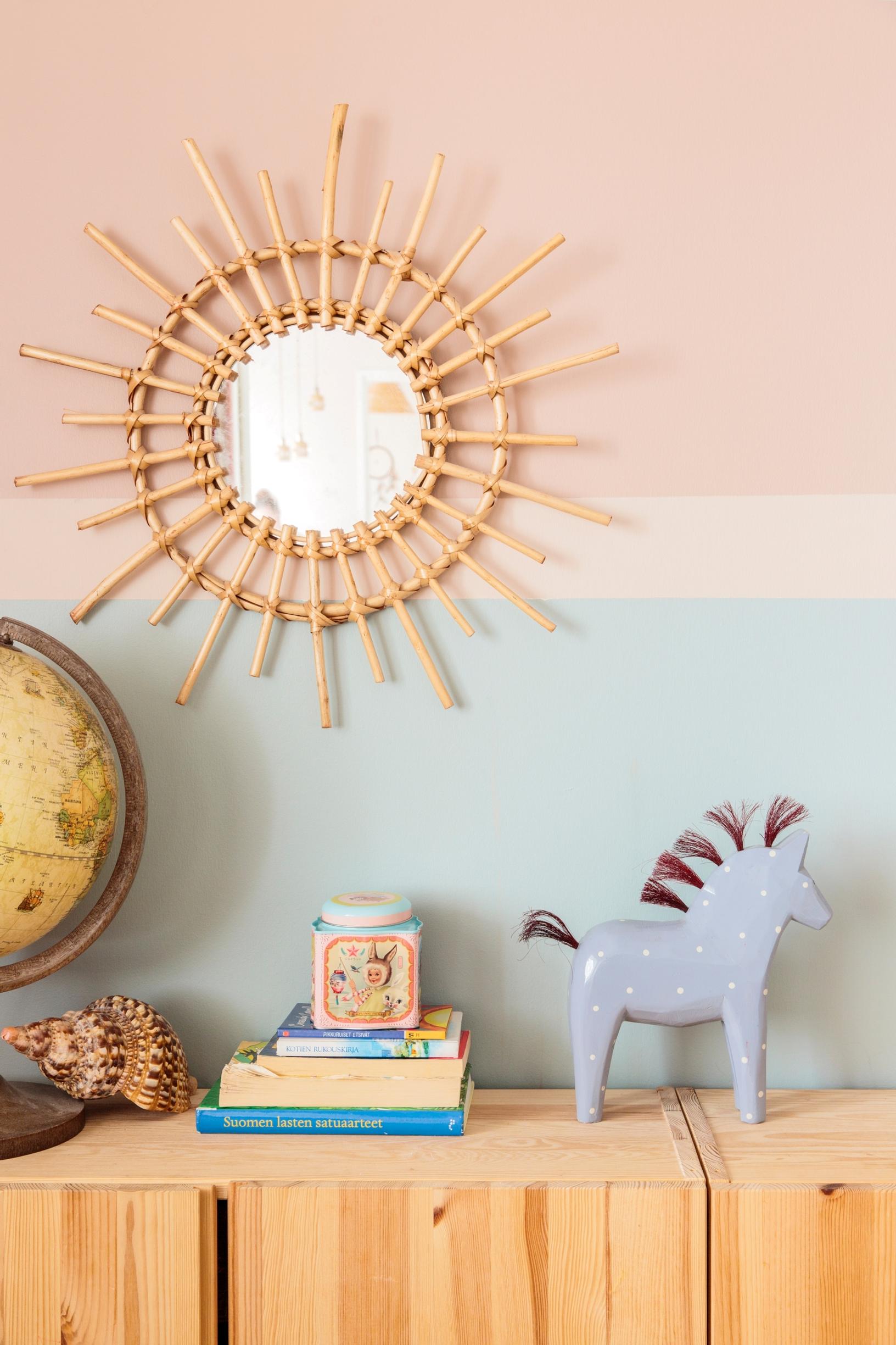 Vaalean puisen lipaston päällä on kasa kirjoja ja puuhevonen. Pöydällä on myös vanha karttapallo ja seinällä roikkuu peili. Seinä on pinkki ja vaaleansininen.