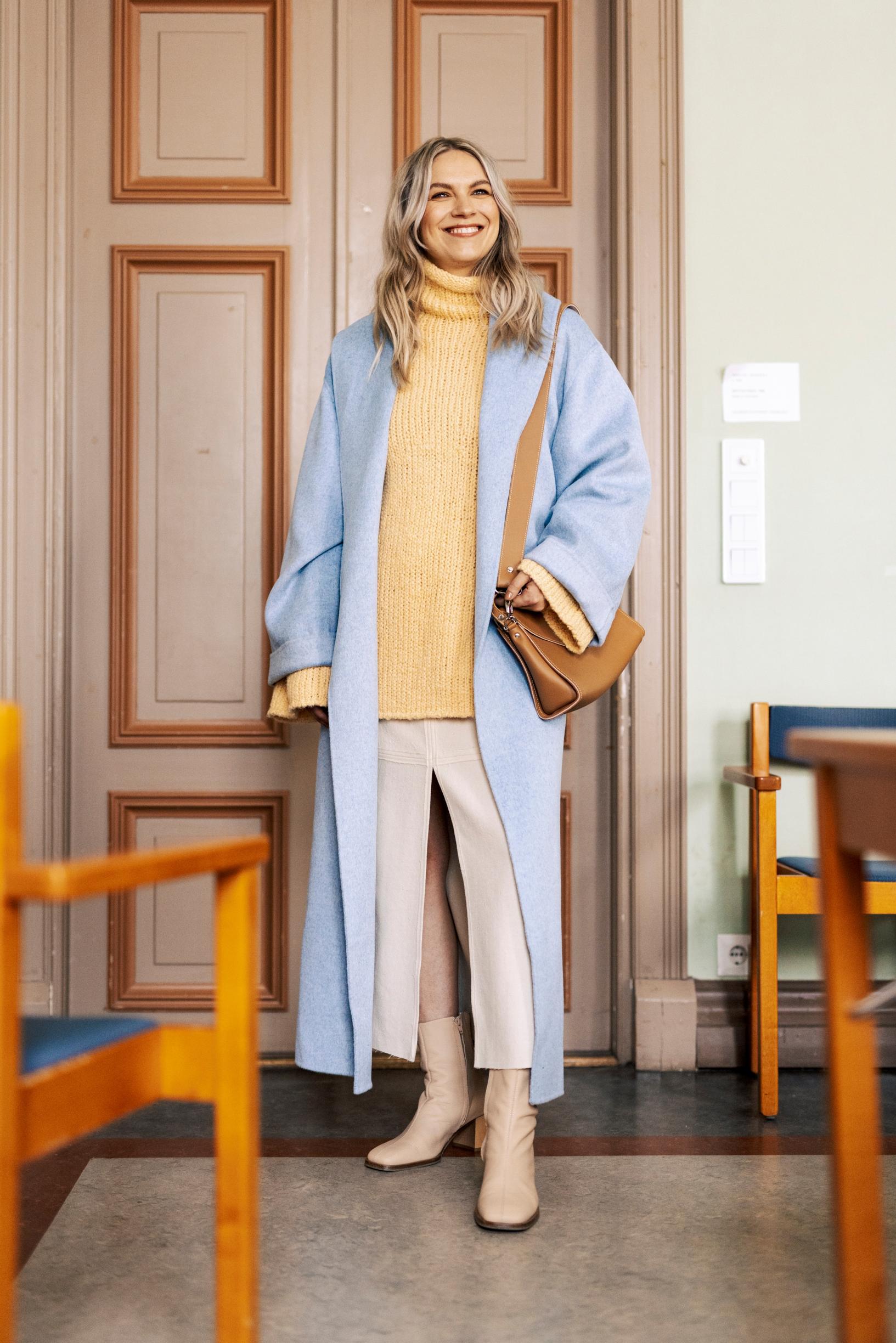 Emmi-Liia Sjöholm on pukeutunut vaaleansiniseen pitkään takkiin ja keltaiseen neuleeseen.